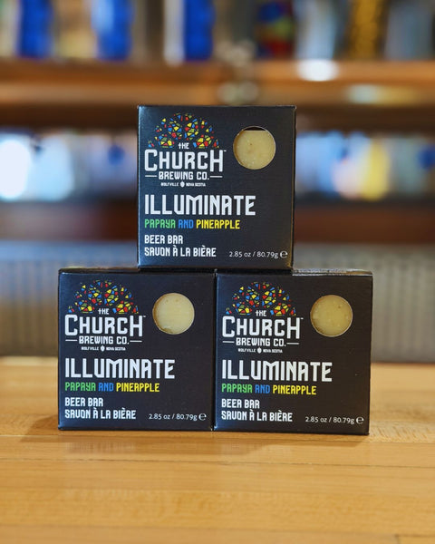 Three boxes of 'The Church Brewing Co. Illuminate' beer bar on a wooden surface with a blurred background.